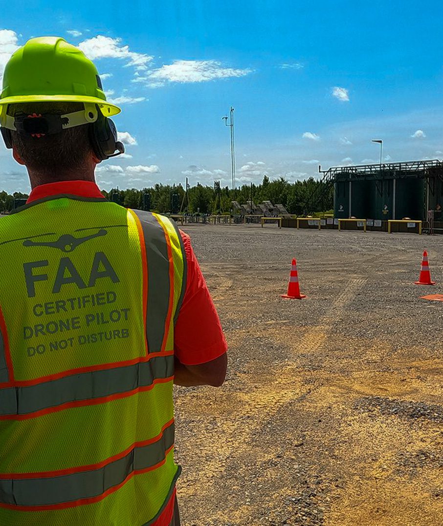 FAA commercial drone pilot, wearing a yellow safety belt, flying over a Natural Gas well pad